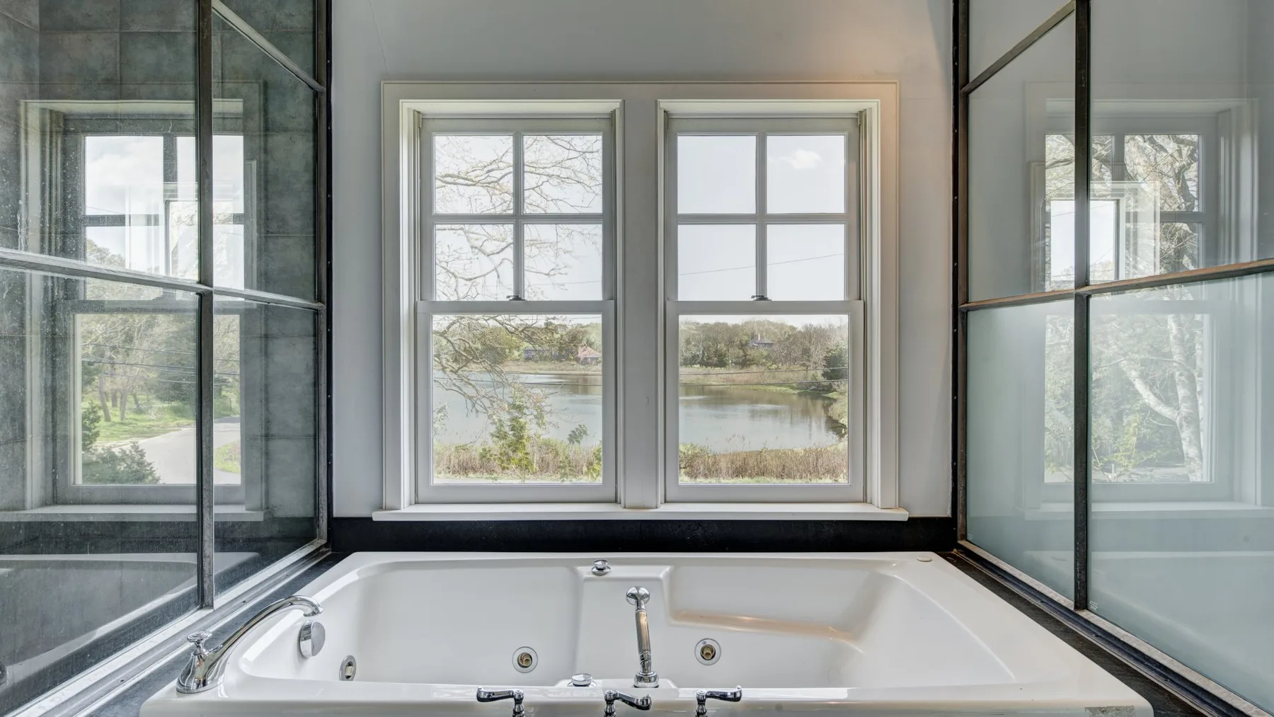 Un baño con una amplia bañera bajo dos ventanas con vistas a un paisaje junto al agua.