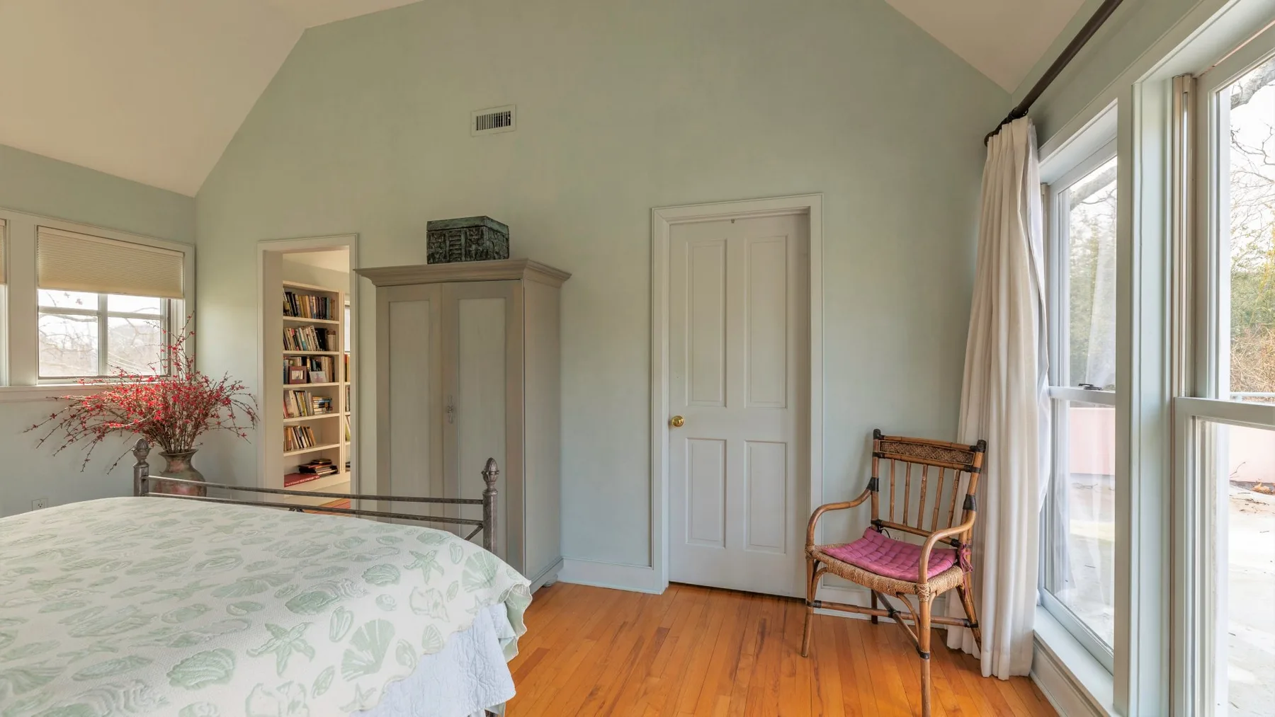 Un dormitorio luminoso con una colcha floral, una silla de madera y grandes ventanas.