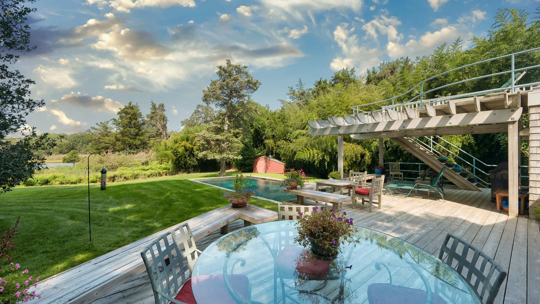 Un patio ajardinado con una mesa de vidrio y asientos al aire libre bajo una terraza elevada.