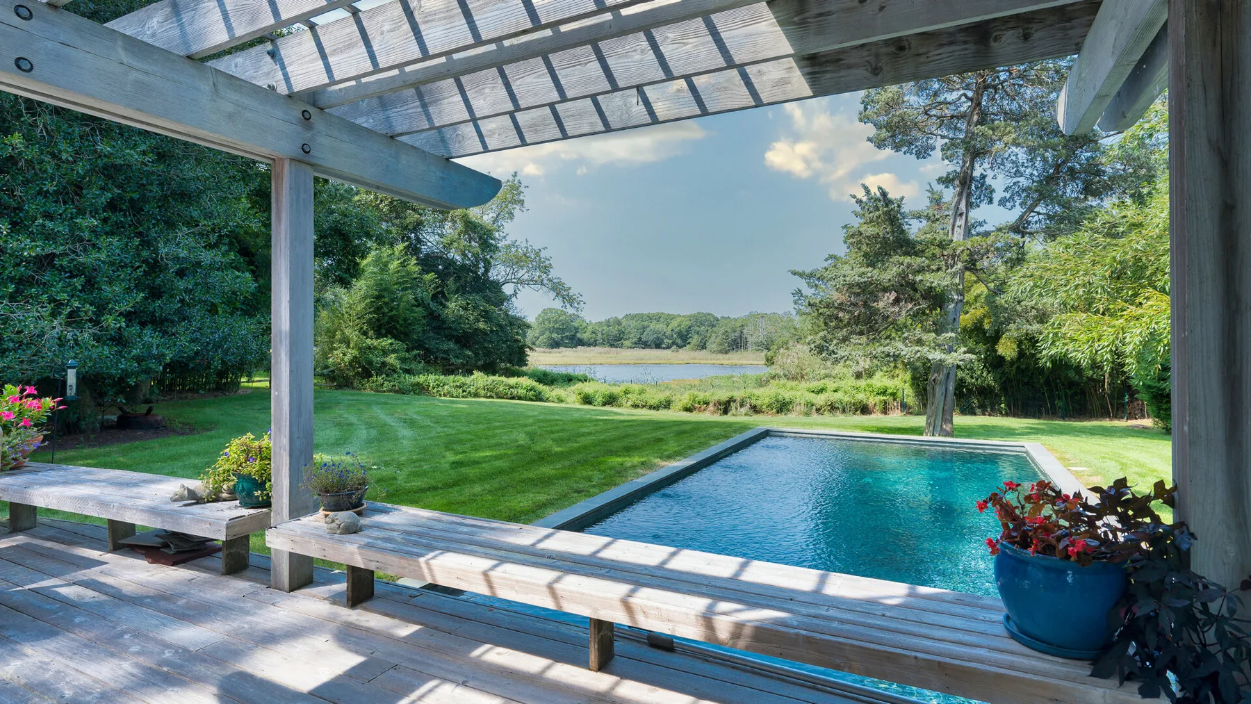Una piscina junto a un césped, rodeada de árboles y una terraza de madera cubierta.