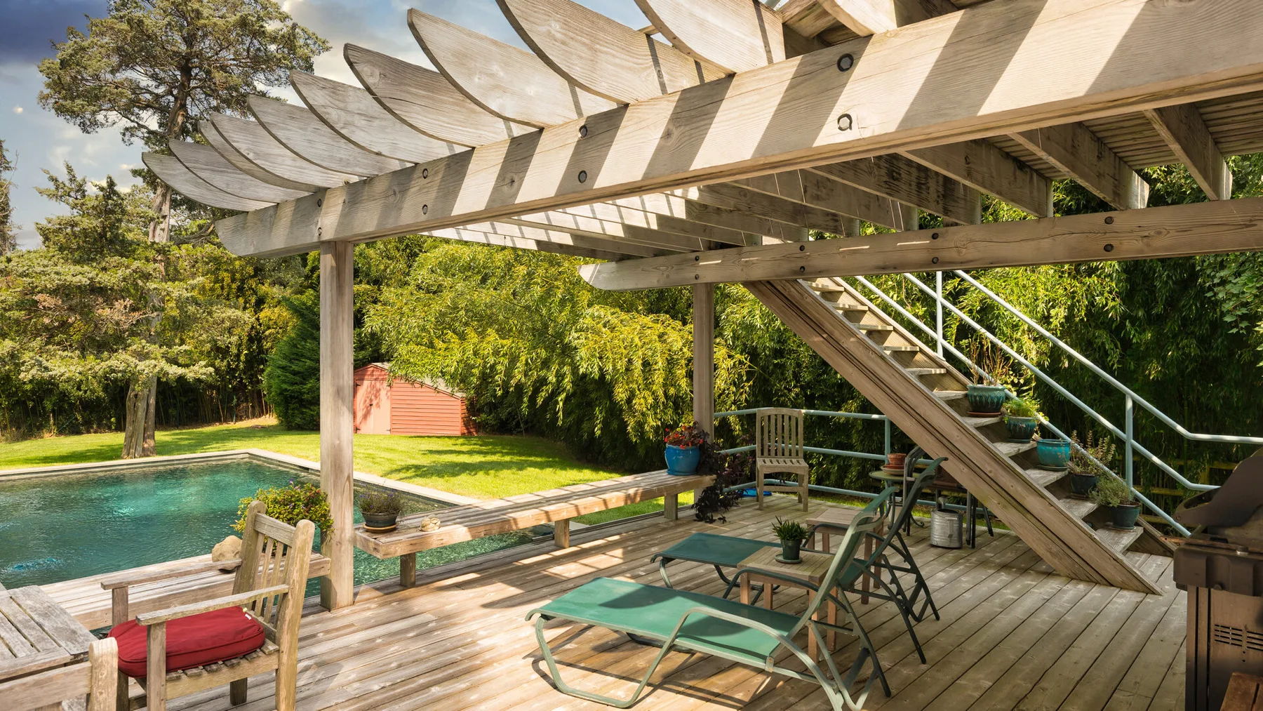 Una terraza de madera junto a la piscina queda bajo un patio elevado con asientos al aire libre.