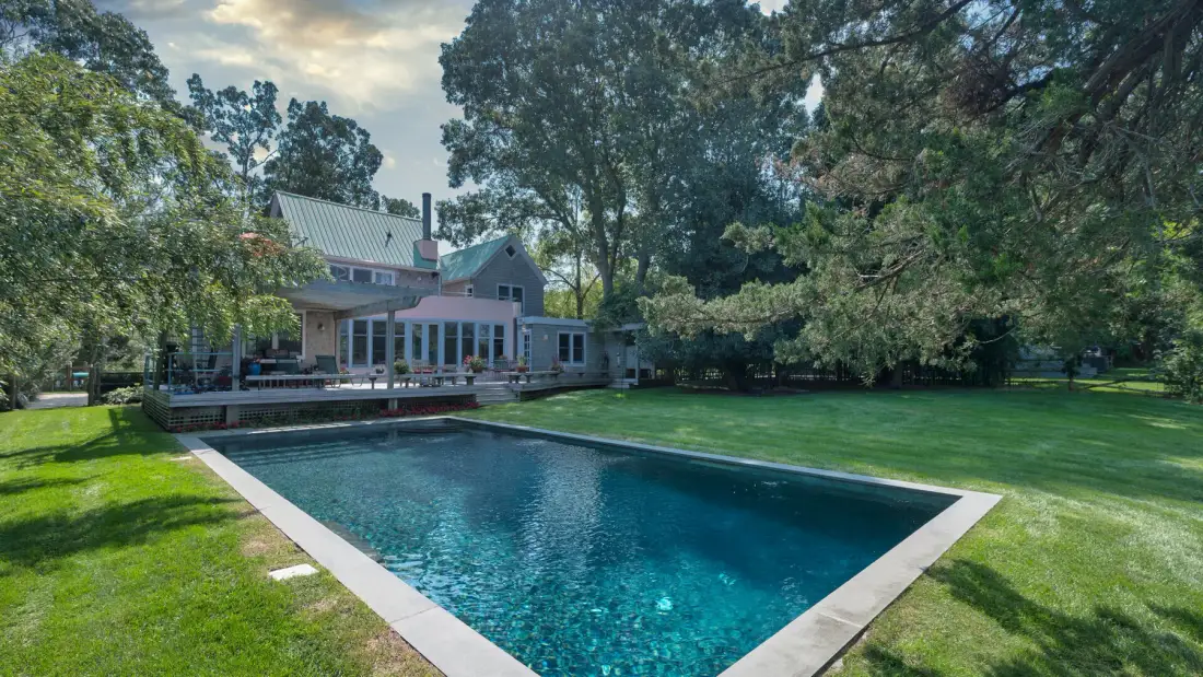 Una piscina junto a una casa, rodeada de césped verde y árboles.