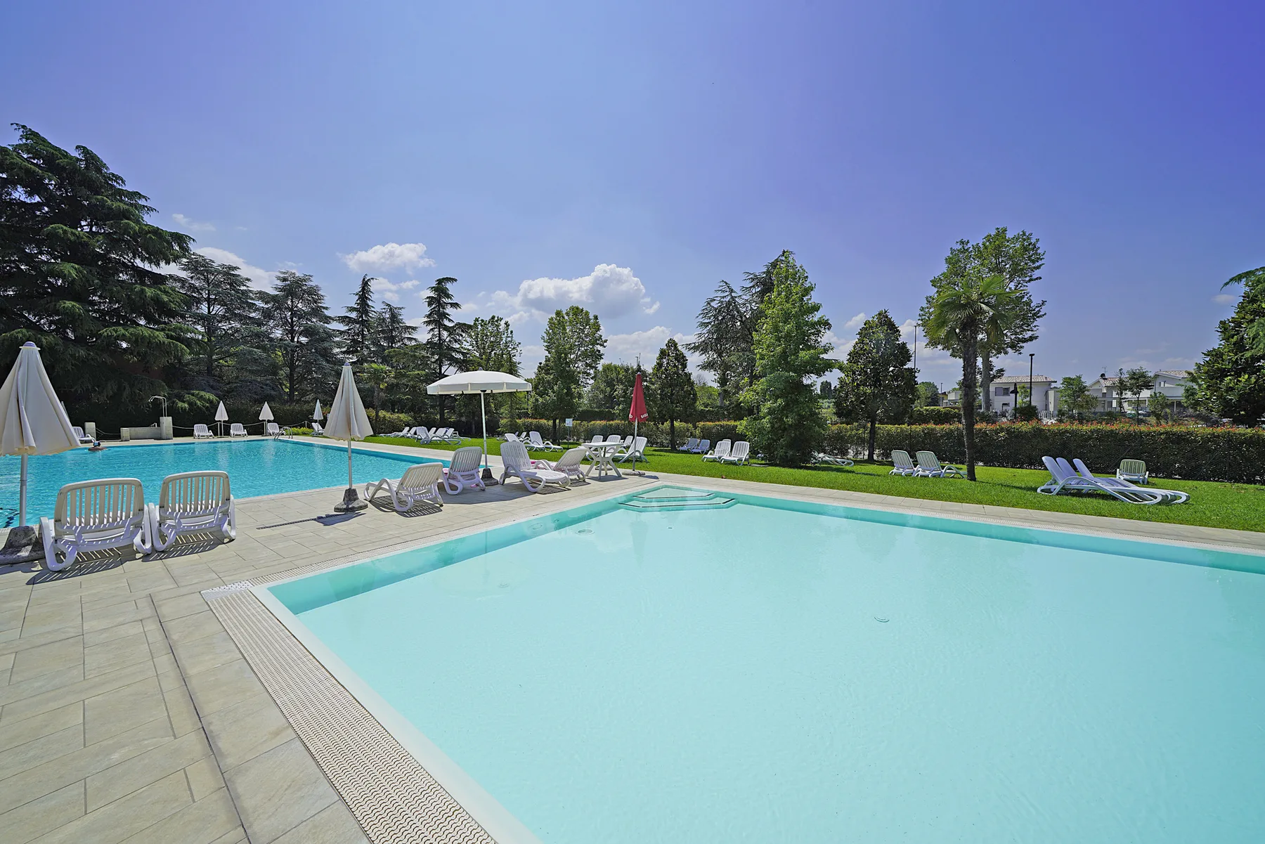 Una piscina al aire libre junto a una terraza pavimentada con tumbonas y árboles.