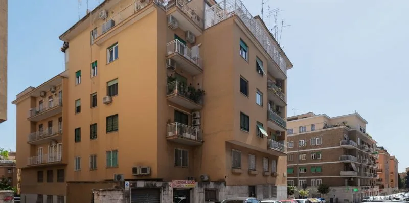 Un edificio de apartamentos color beige con balcones se alza junto a una calle urbana.