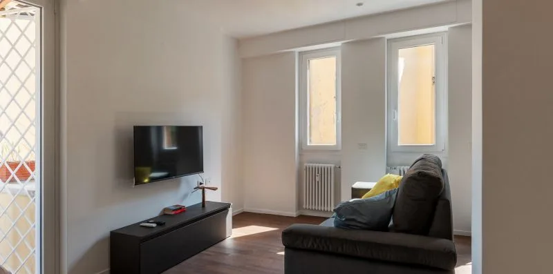 Una sala de estar con televisor, mueble bajo y asientos oscuros junto a ventanas altas.
