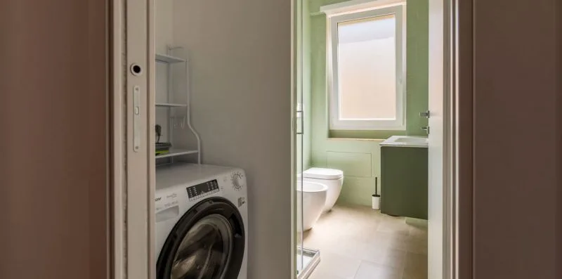 Un baño junto a una lavadora, con una ventana sobre el inodoro.