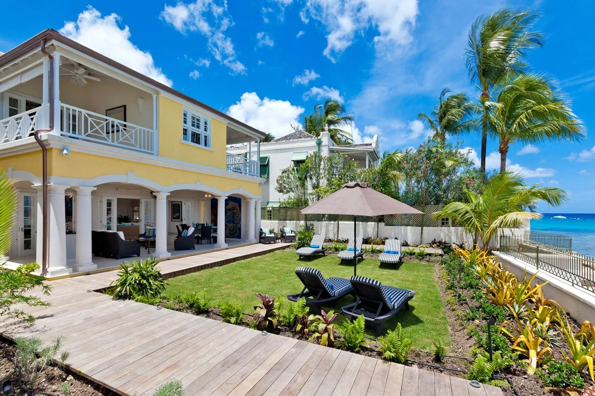 A seaside house and garden with a lawn, patio seating, and palm trees.