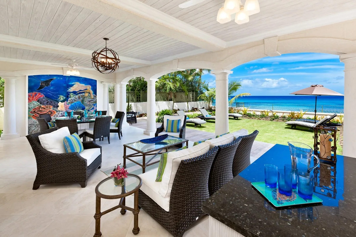 A covered seaside patio with outdoor seating overlooks a lawn and the ocean.