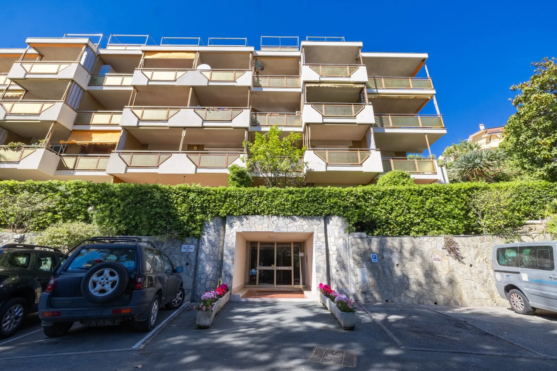 An apartment building entrance is framed by a stone wall and greenery.
