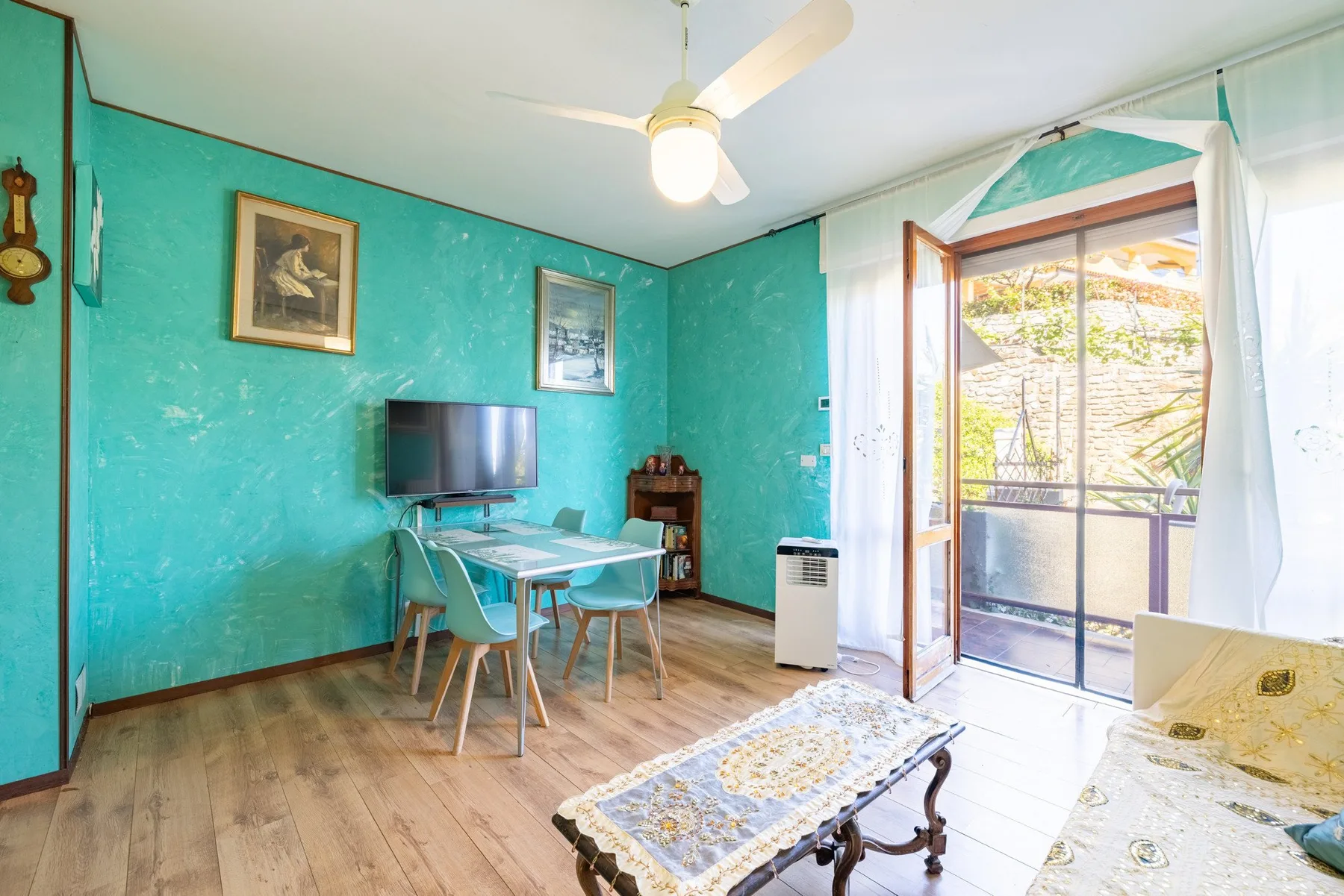 A turquoise living area has a dining table and an open balcony door.