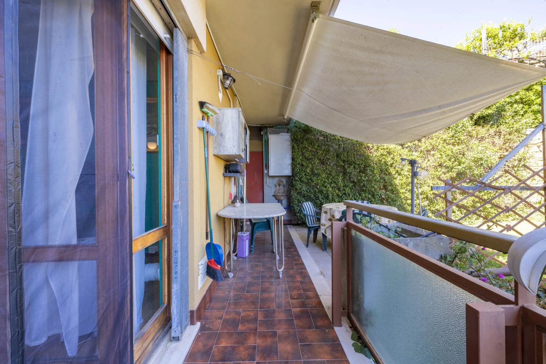 A narrow outdoor balcony with tiled flooring, a table, and a retractable shade.