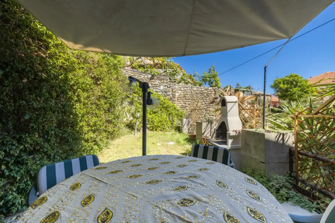 An outdoor dining area sits beneath a shade canopy beside a stone barbecue.