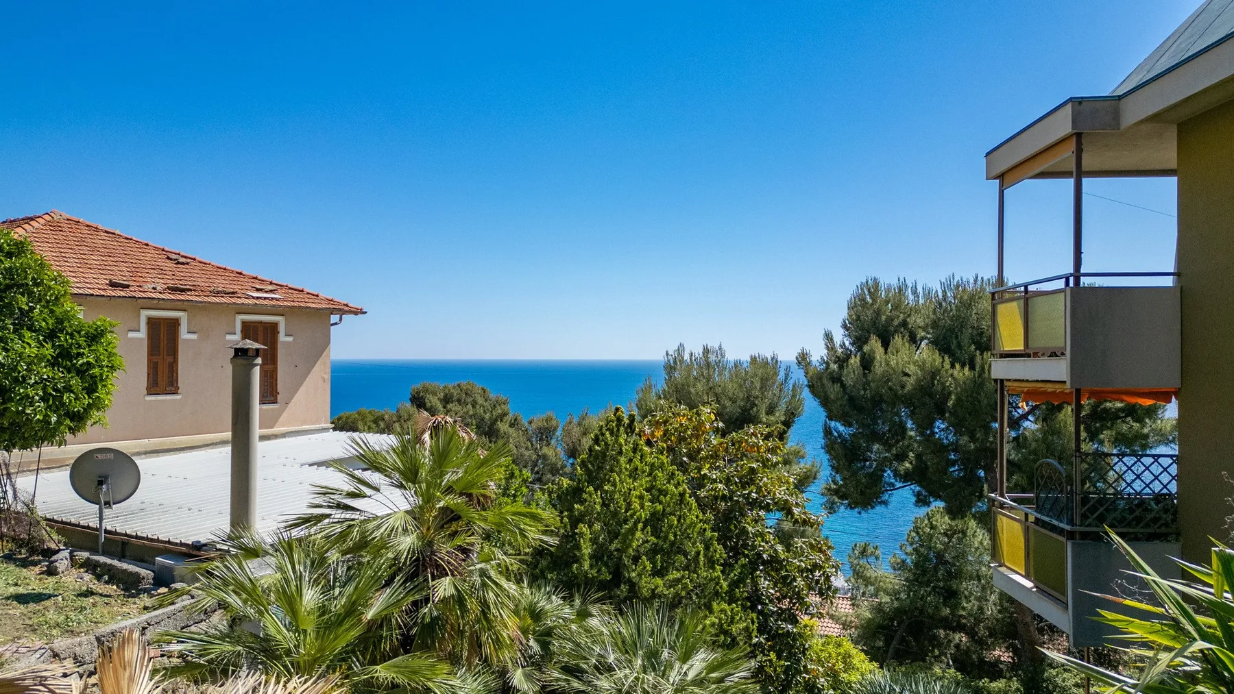A coastal view shows the sea between leafy trees and nearby buildings.