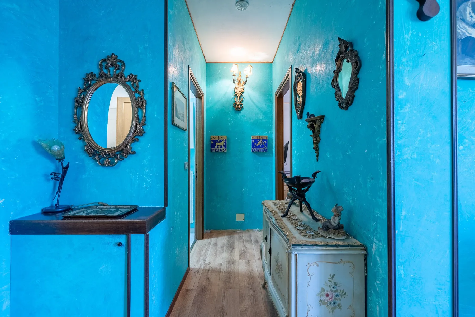 A turquoise hallway with decorative mirrors, wall art, and a narrow console.