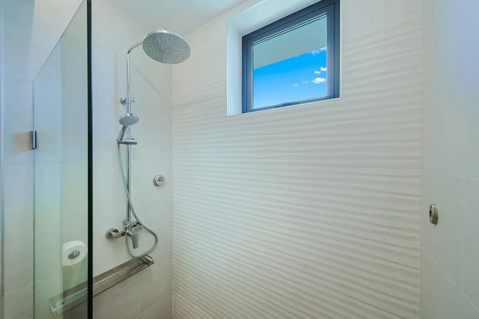 Your private spa-like bathroom features a rainfall shower and handheld option. Light pours through the window as you refresh after exploring, wrapped in serene white tile and gleaming fixtures.