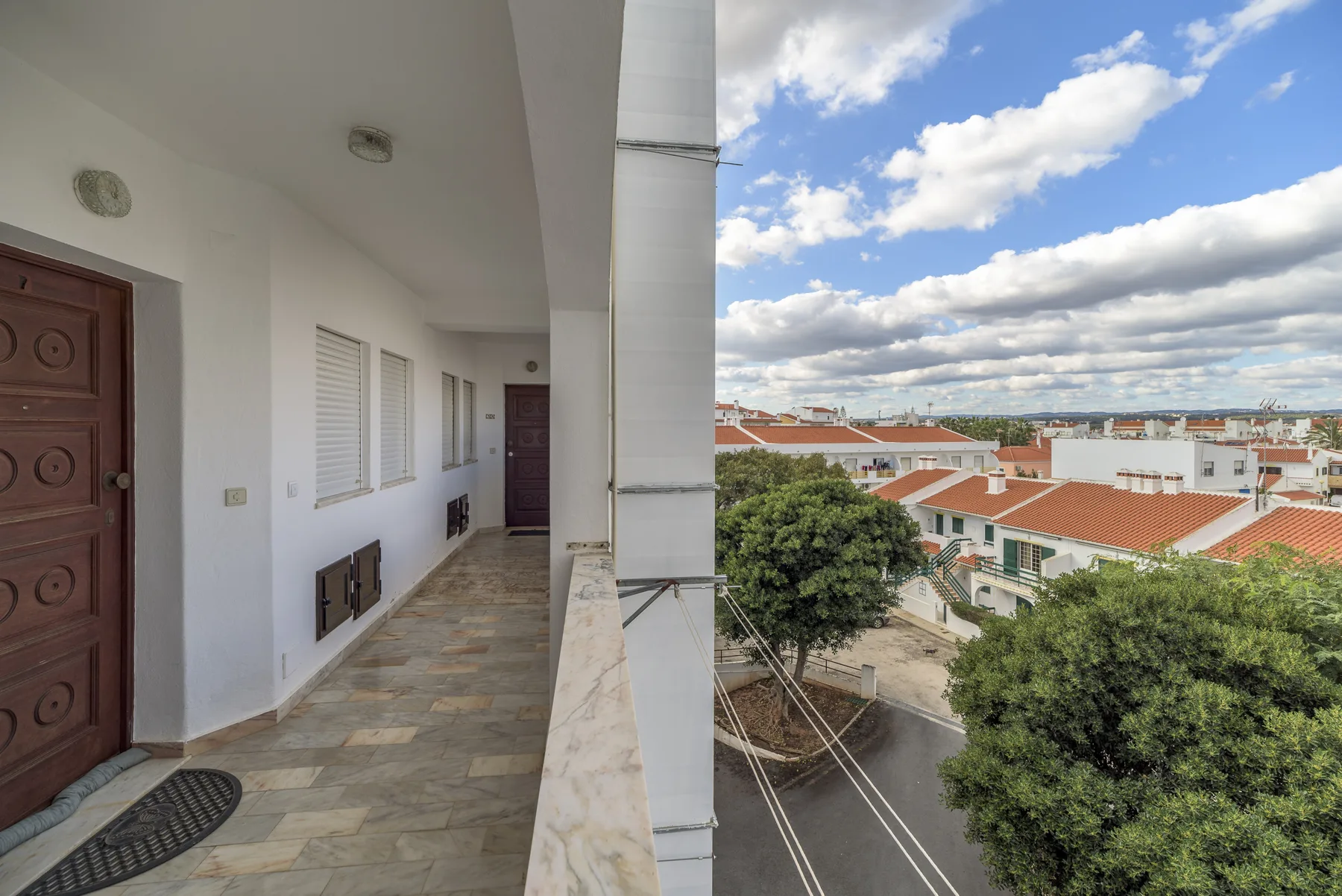 El pasillo del balcón del apartamento da a edificios cercanos y árboles frondosos bajo un cielo parcialmente nublado.