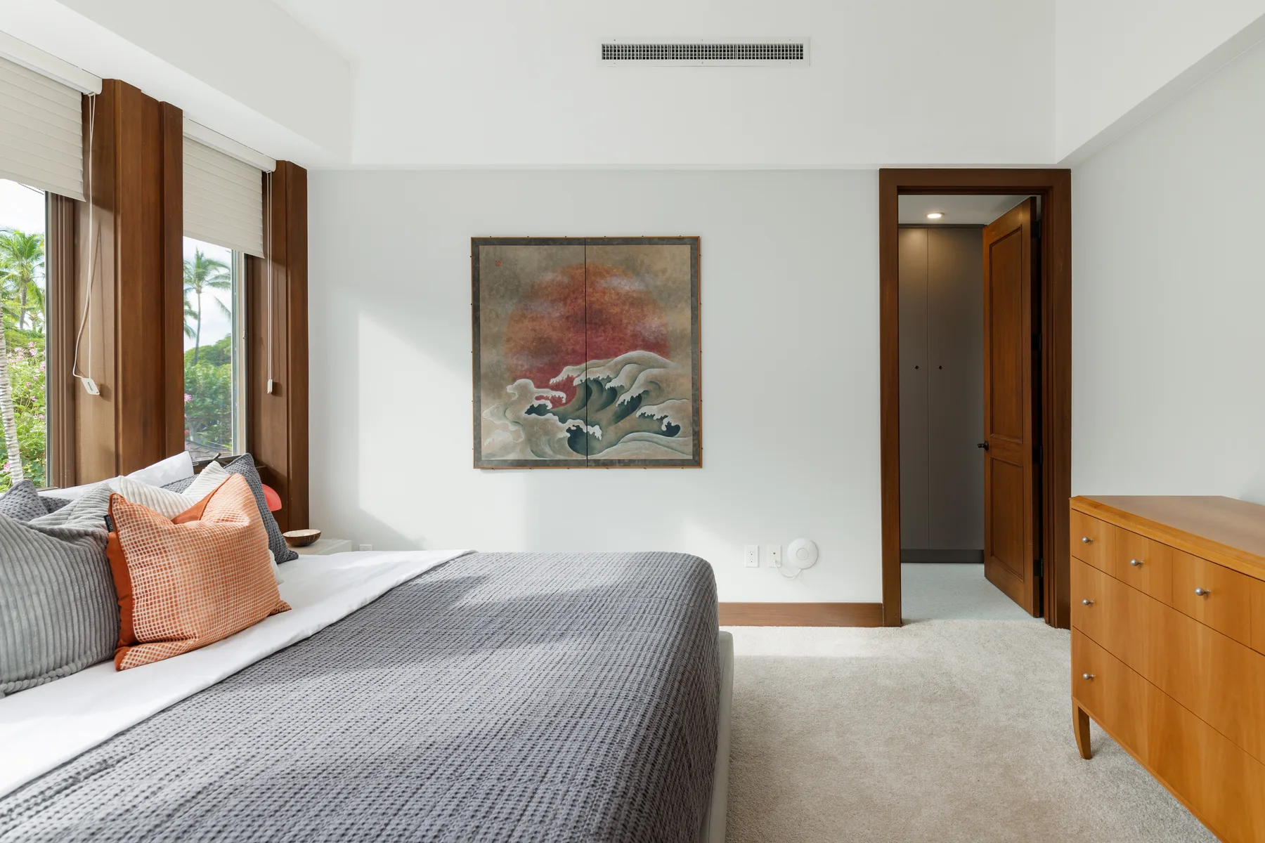 Un dormitorio luminoso con una cama junto a ventanas altas y un cuadro en la pared.
