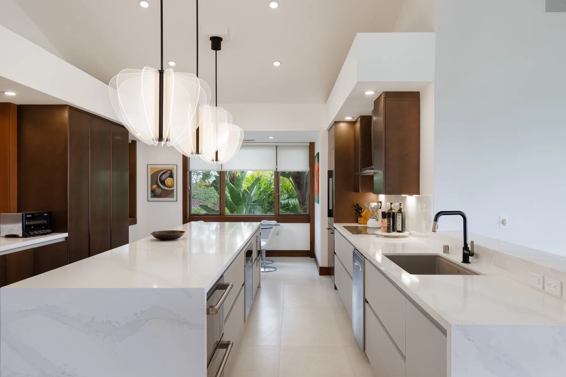 Una cocina moderna y luminosa con encimeras blancas, gabinetes oscuros y lámparas colgantes.