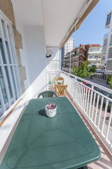 Un balcón estrecho tiene una mesa verde, asientos y vistas de edificios cercanos.