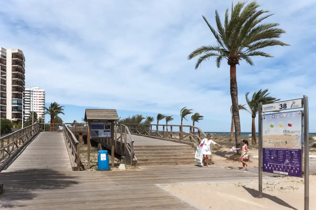 Una pasarela junto a la playa pasa junto a un refugio de madera, con palmeras y el mar al fondo.