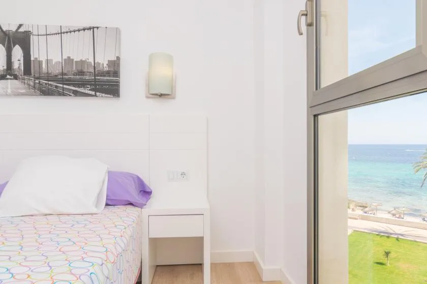 A bedroom with a bed beside a large window overlooking the sea.