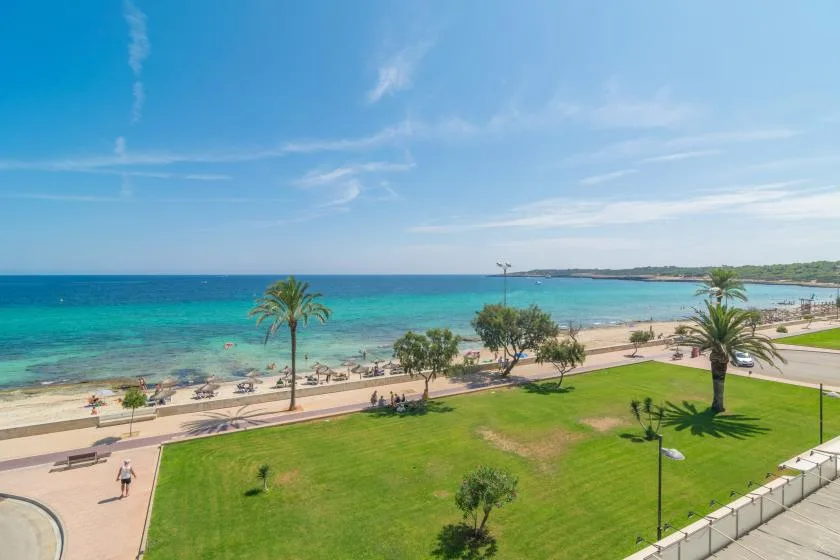 A seaside promenade borders a green lawn, with turquoise water and a sandy beach beyond.
