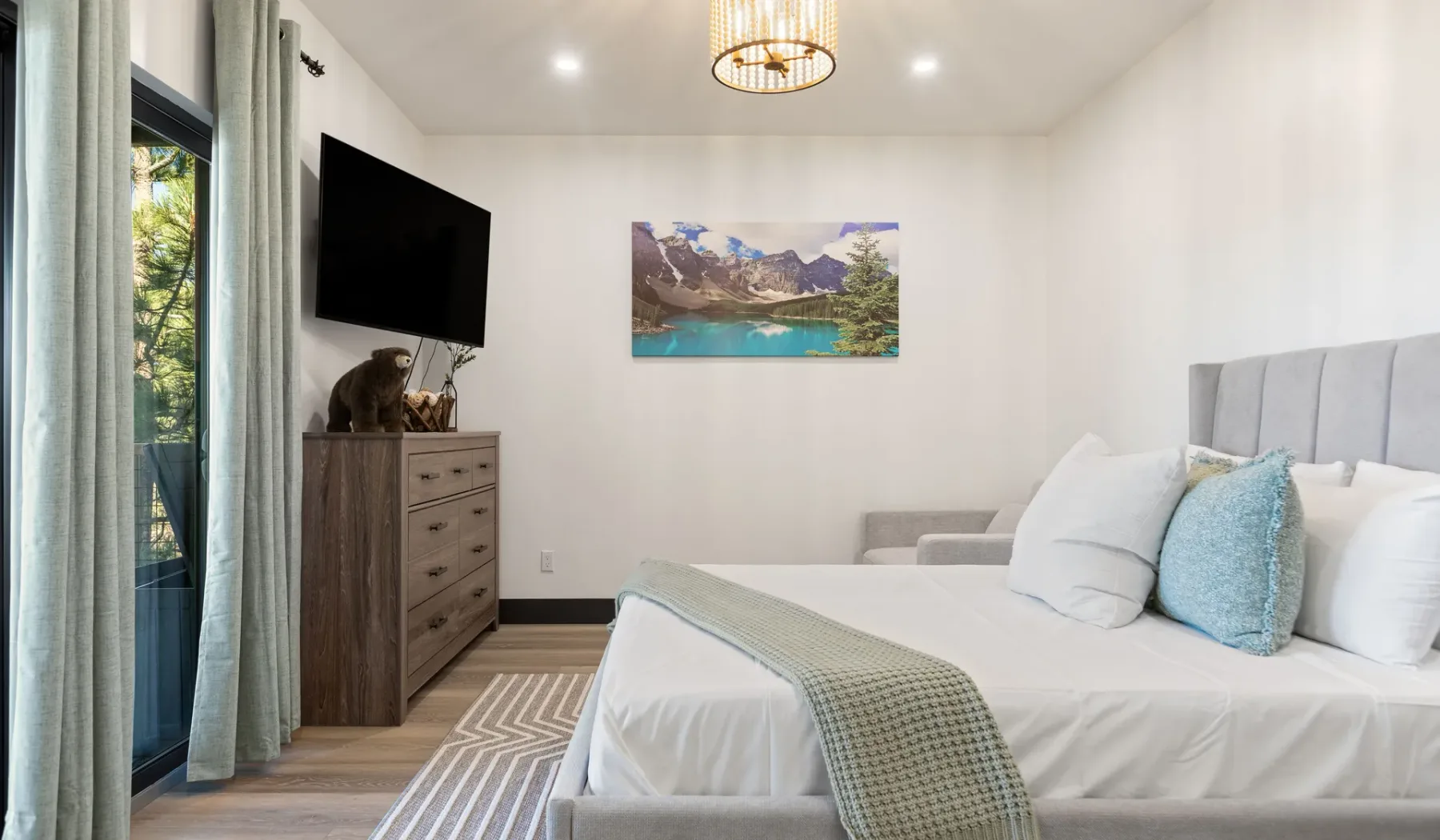 Un dormitorio luminoso con una cama bien arreglada, cortinas claras y un televisor en la pared.