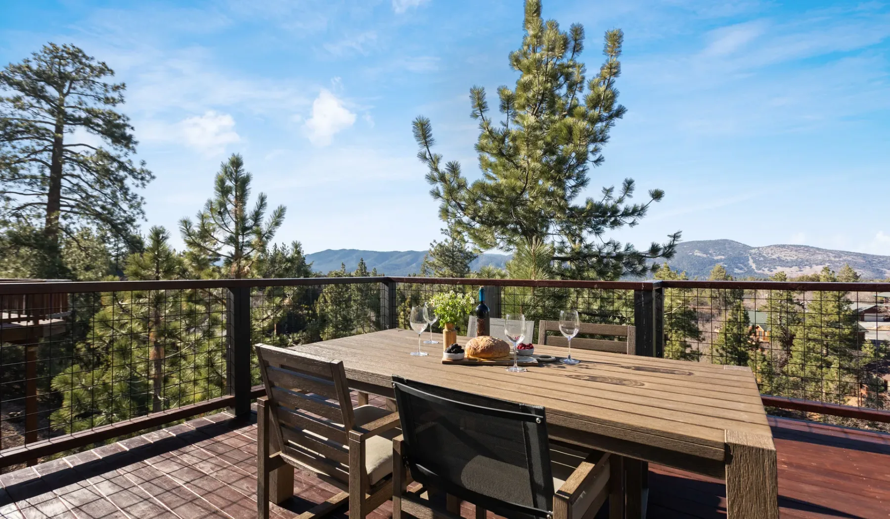Una terraza al aire libre tiene una mesa de comedor de madera y sillas, con vistas a árboles y montañas lejanas.