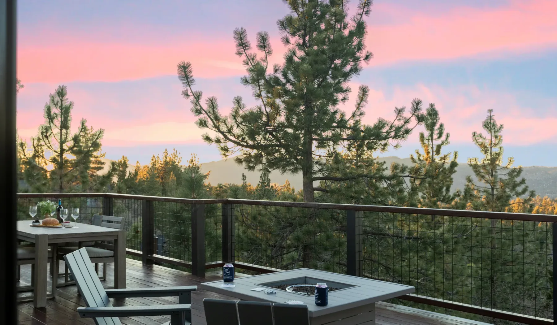 Una terraza exterior con mesas y asientos da a pinos bajo un cielo rosado al atardecer.