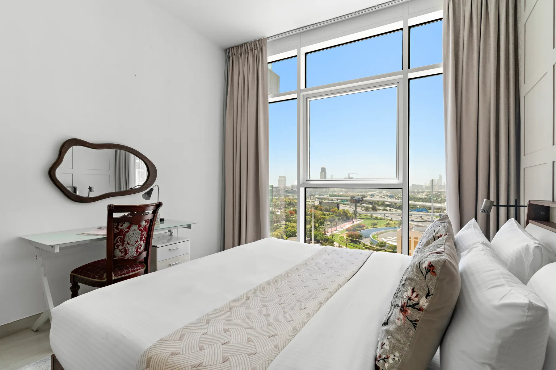 A bright bedroom with a bed beside large windows overlooking the city.