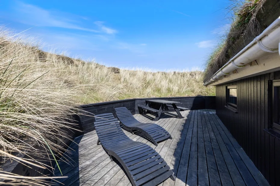 Una terraza de madera con asientos reclinables y una mesa de picnic está junto a hierba alta de dunas.