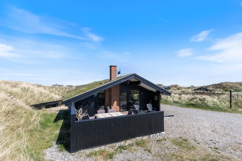 Una cabaña de madera oscura se encuentra junto a dunas de arena bajo un cielo azul despejado.