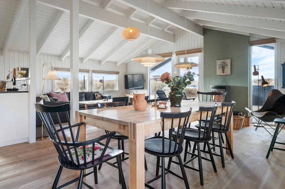Comedor con mesa de madera, asientos oscuros y grandes ventanas.