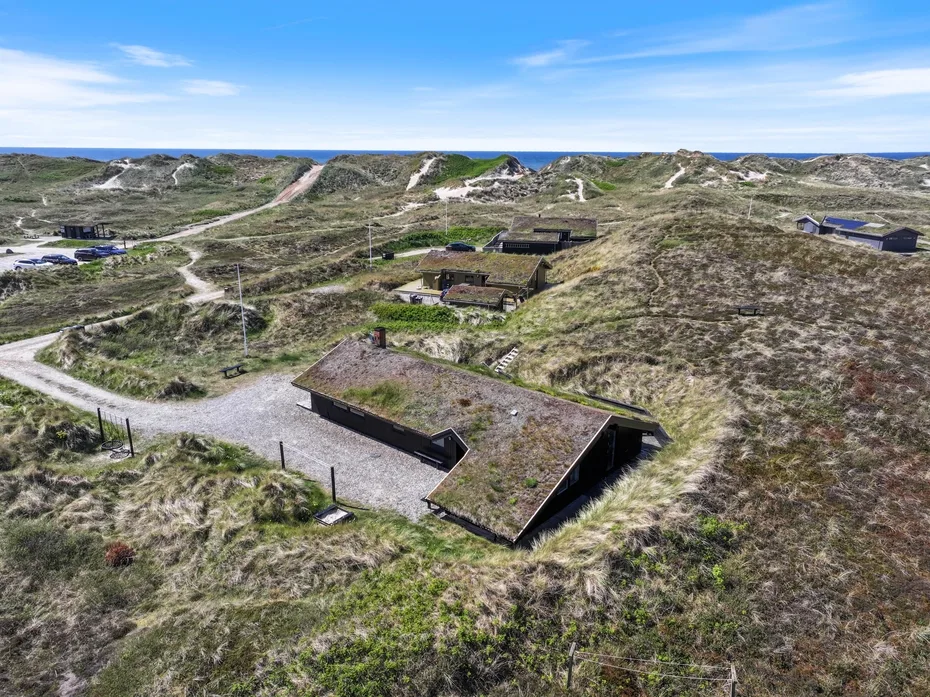 Vista aérea de edificios bajos entre dunas cubiertas de hierba, con el mar en el horizonte.