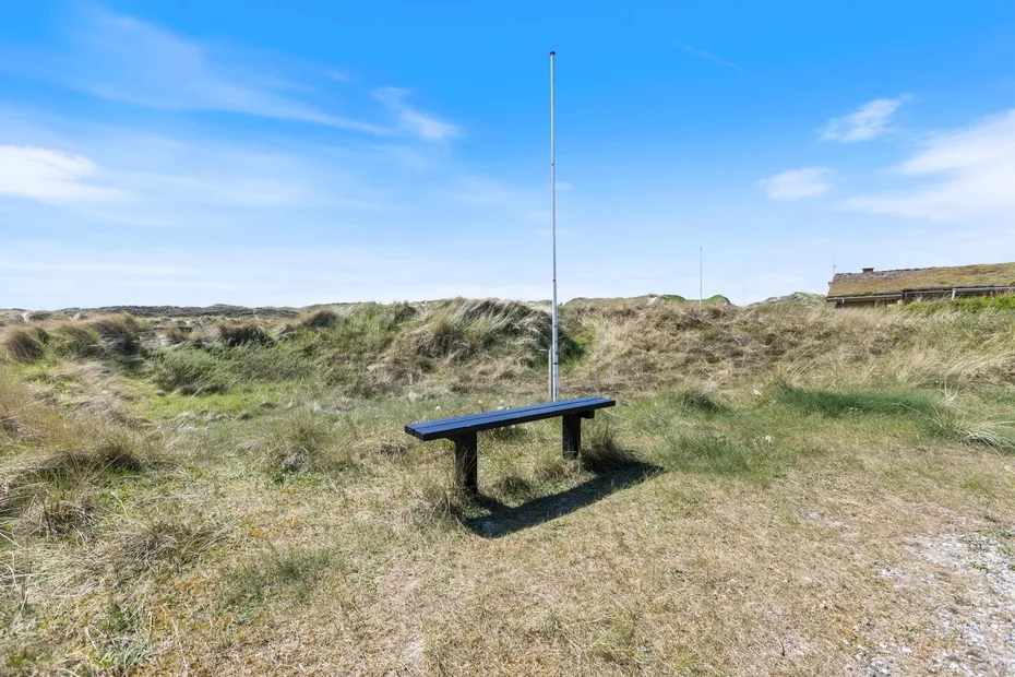 Un pequeño banco exterior está en un paisaje de dunas cubiertas de hierba bajo un cielo despejado.