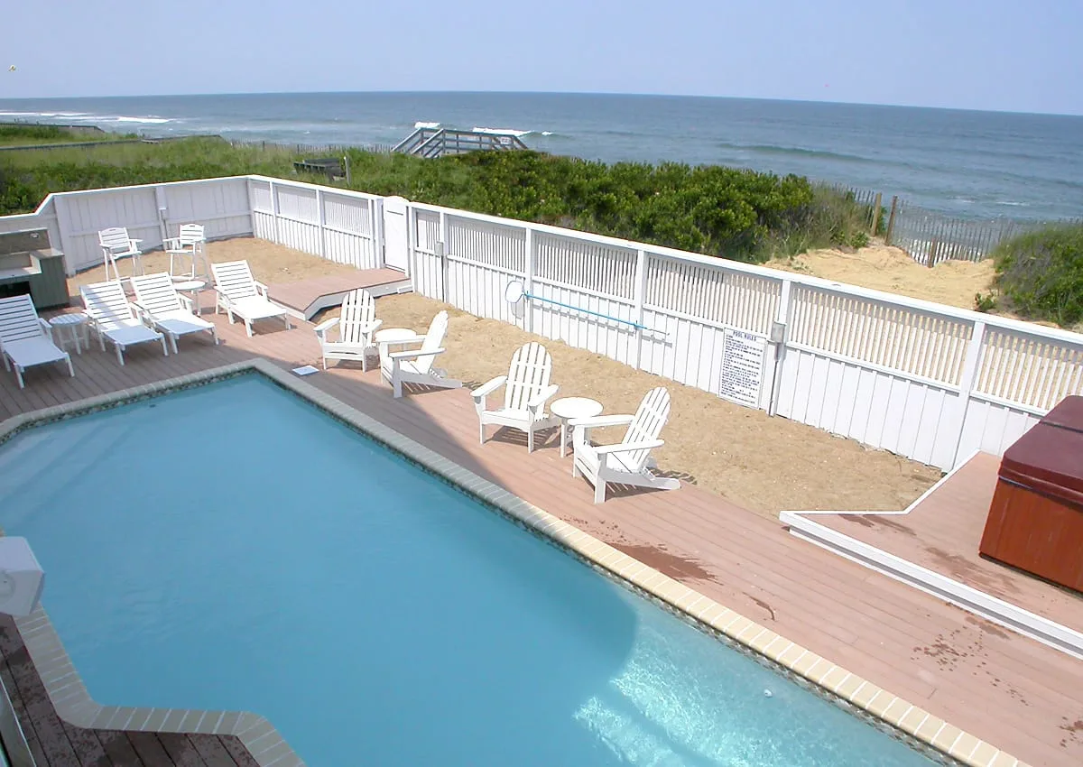 Piscina al aire libre junto a una terraza de madera, con asientos y una playa al otro lado de la cerca.