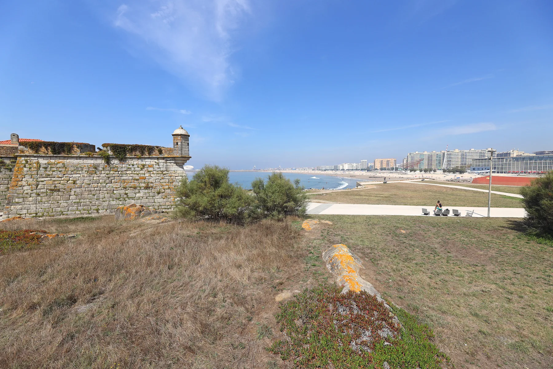 A coastal landscape with a stone fortification, beach, and distant buildings.