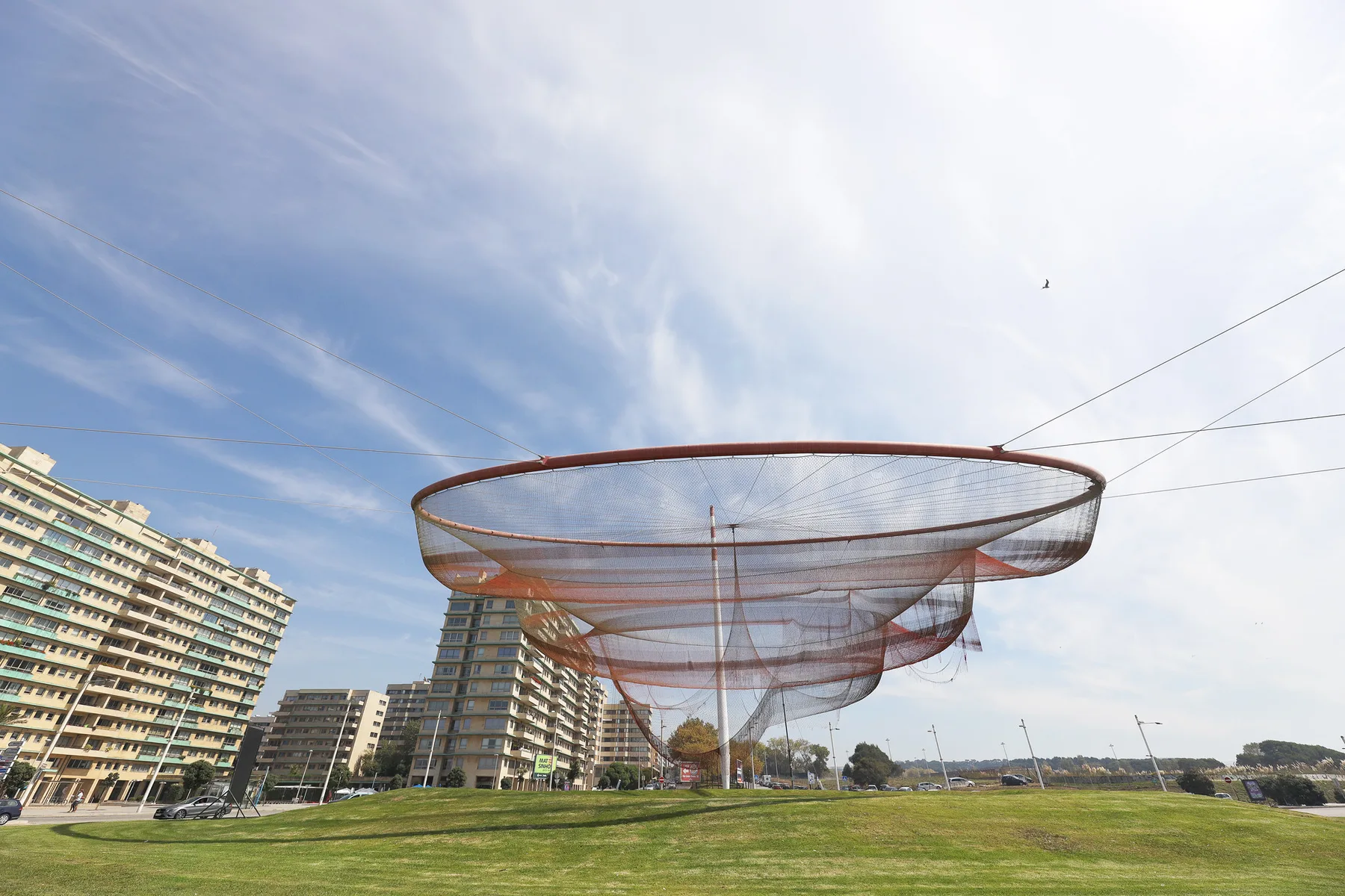 A large net sculpture stretches above a grassy lawn beside modern apartment buildings.