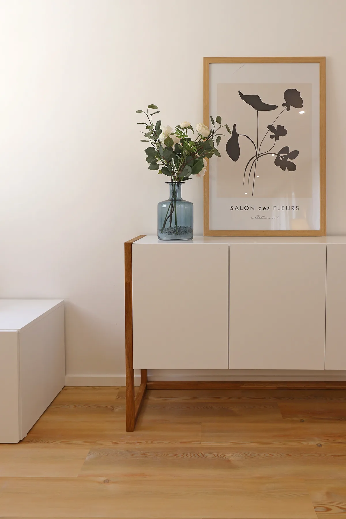 A bright room features a wooden sideboard topped with a vase of flowers and framed artwork.