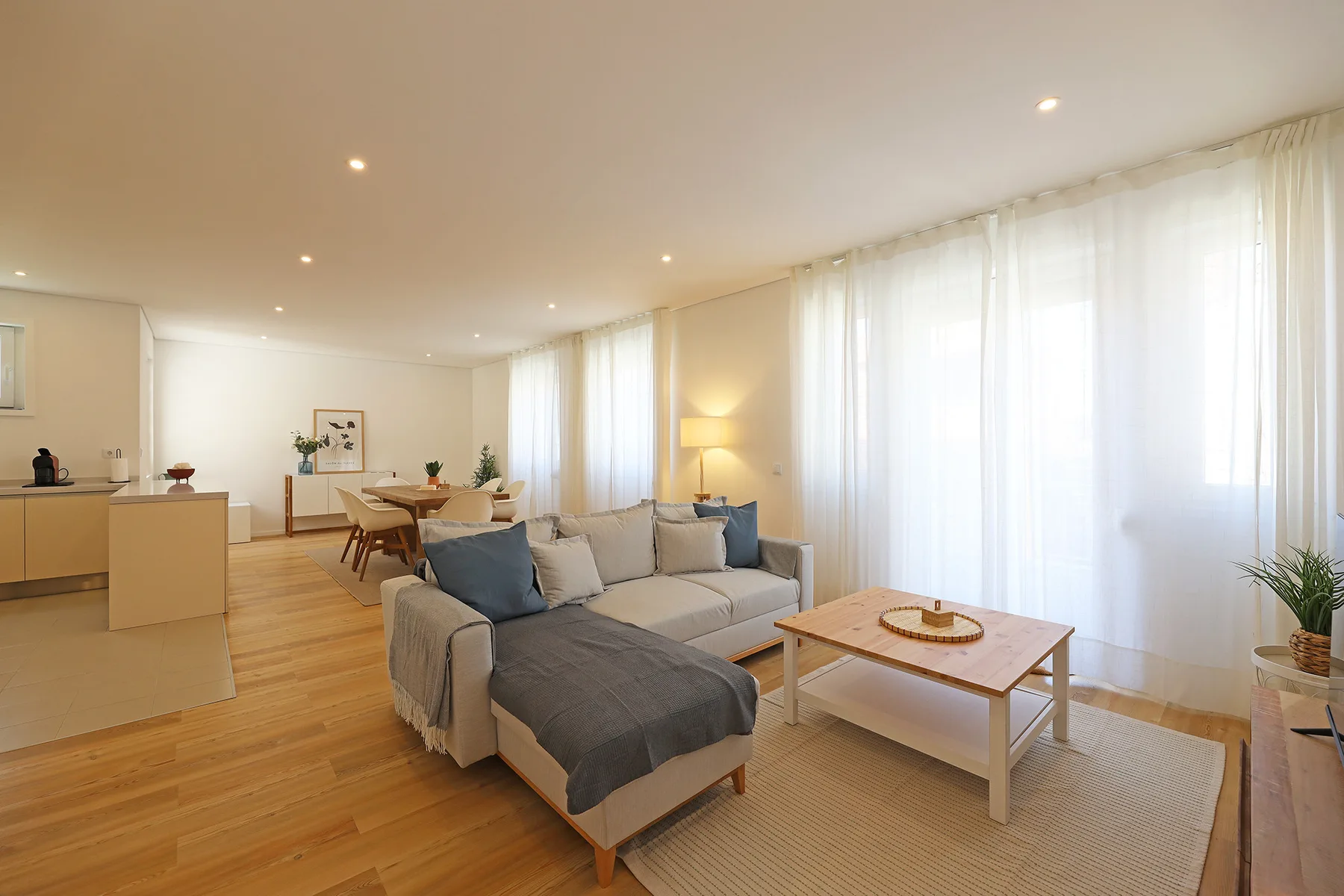 A bright living and dining area has seating, a coffee table, and tall curtained windows.