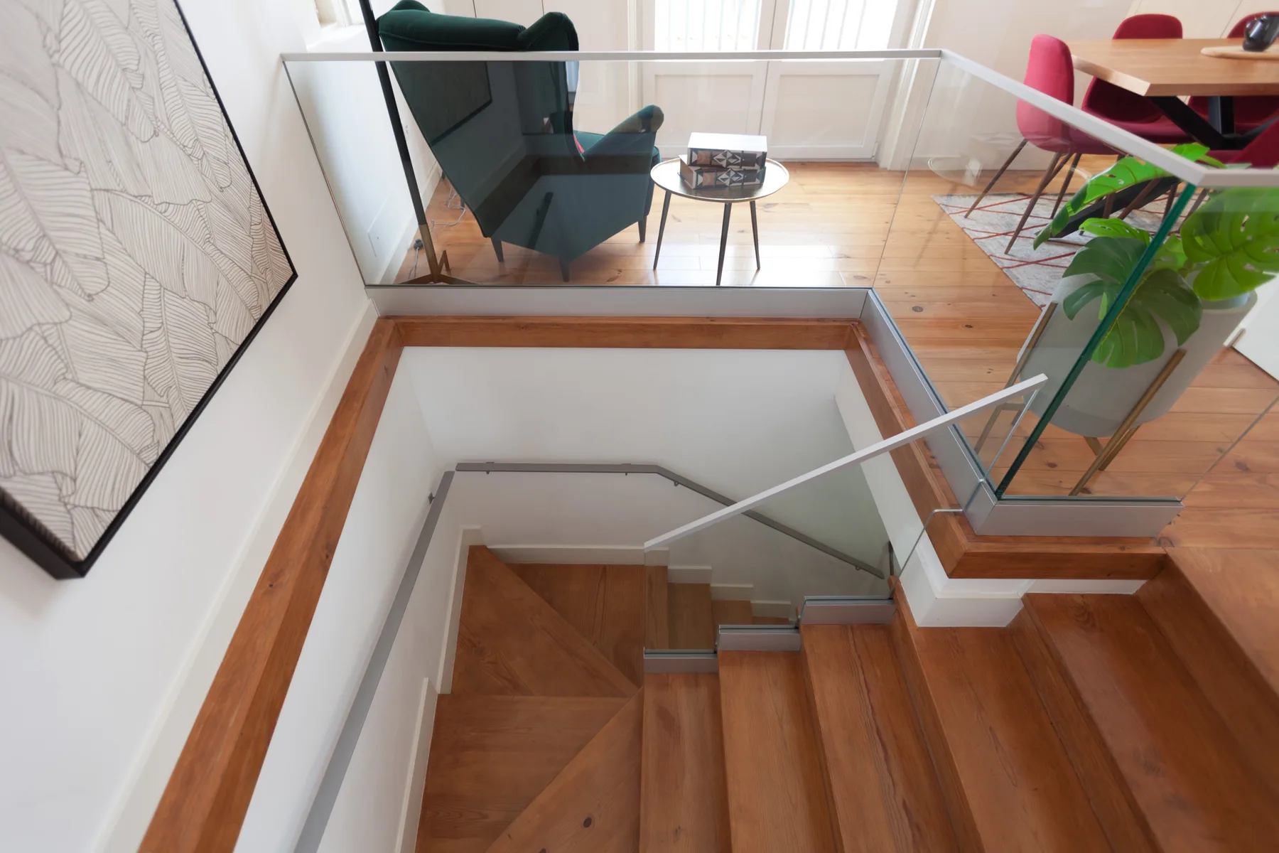 A wooden staircase descends beside a glass railing, with seating and a dining area above.
