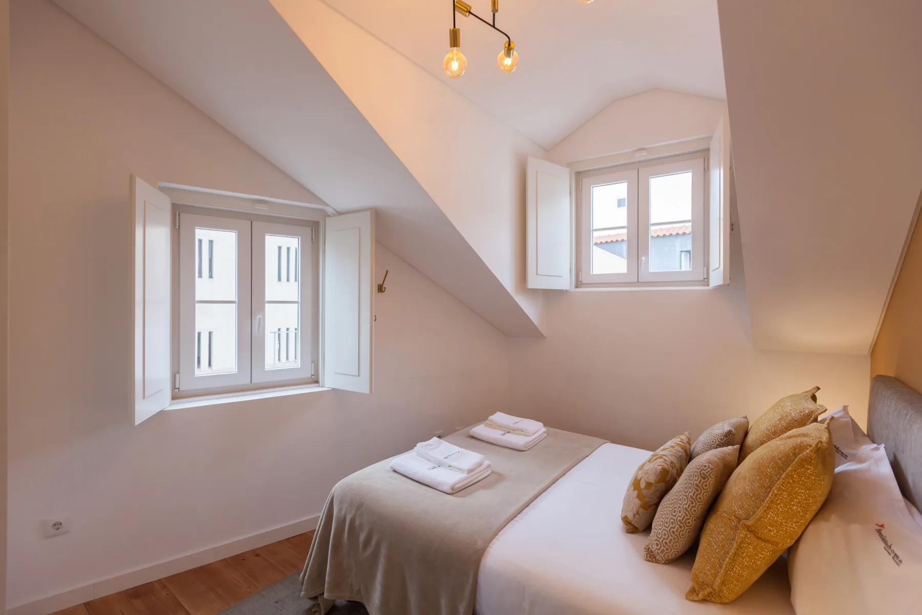 A bedroom with a made bed beneath sloped ceilings and two windows.