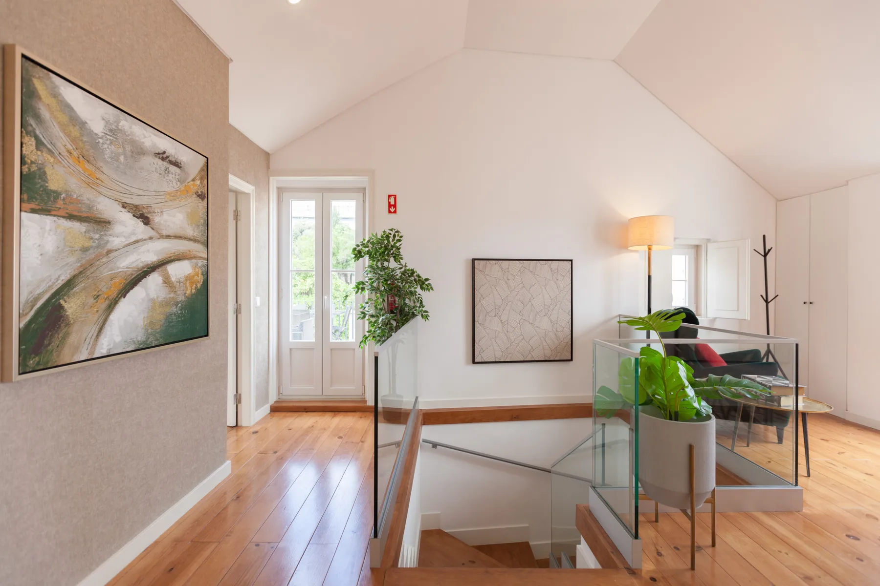 A bright upper-floor living area has glass railing, leafy plants, and framed artwork.