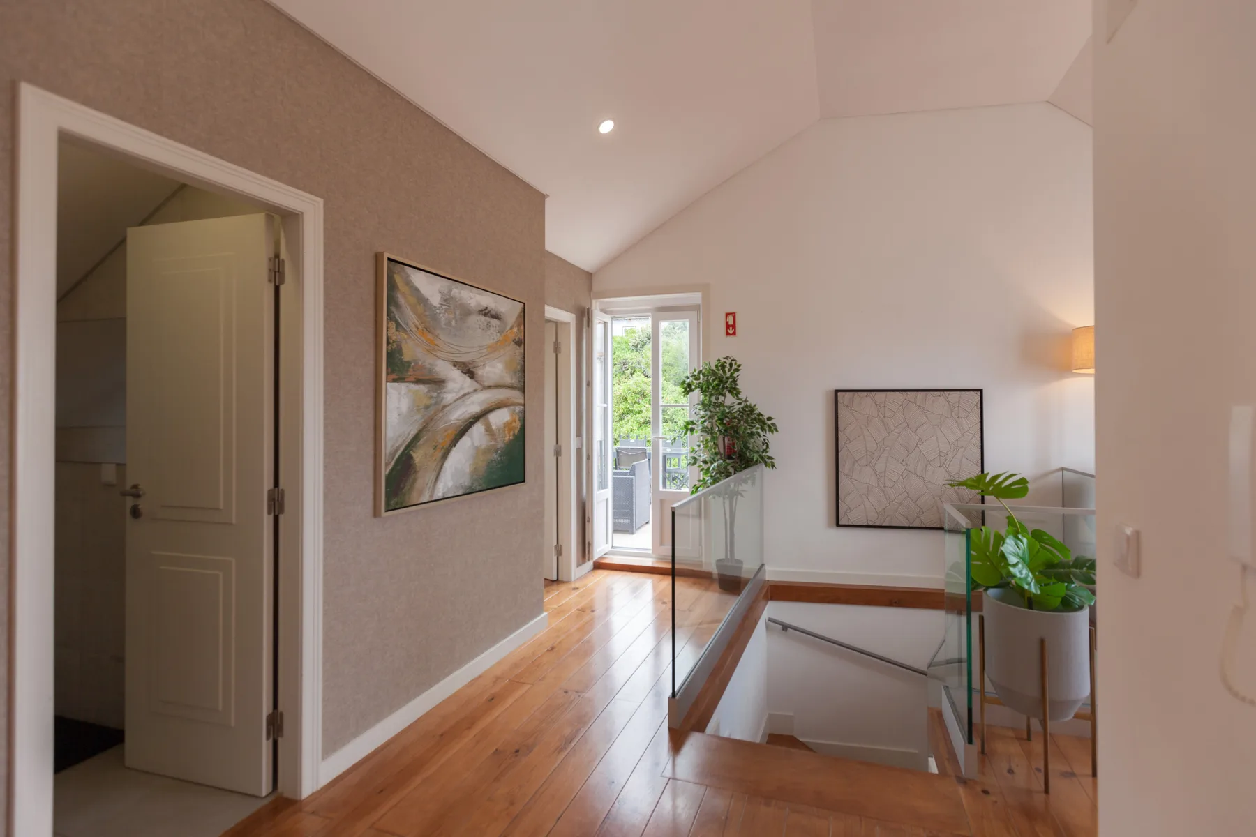 A bright upstairs hallway opens onto a stairwell, with artwork and plants along the glass railing.