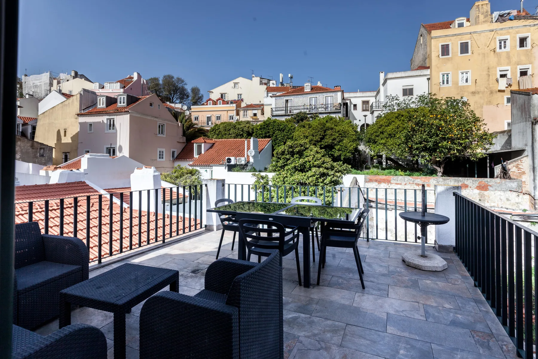 A rooftop terrace with outdoor seating overlooks nearby buildings and trees.