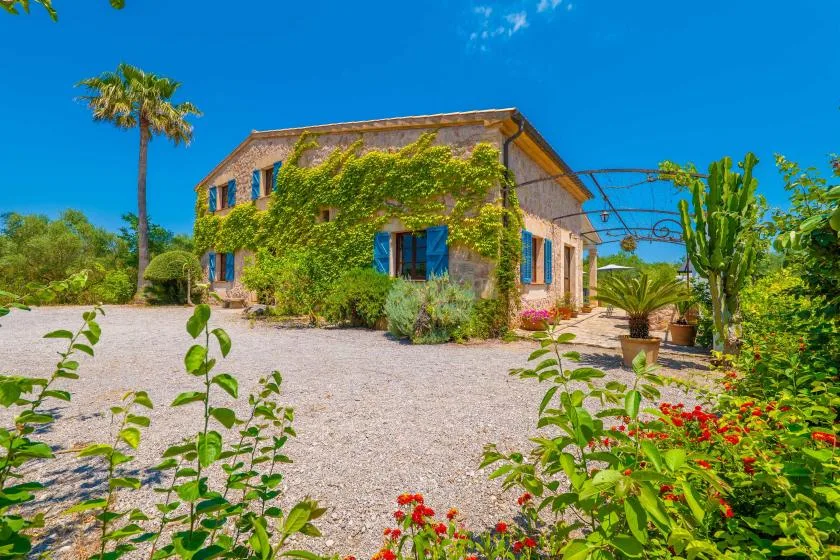 A vine-covered house stands beside a gravel drive amid flowering plants.