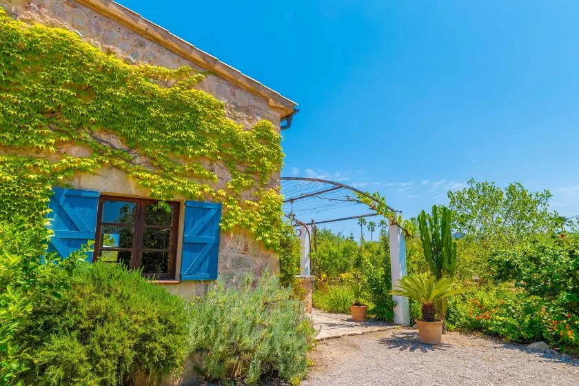 A stone building with blue shutters stands beside a garden path and leafy greenery.