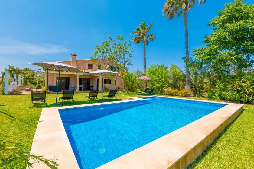 A rectangular swimming pool sits on a lawn beside a house and shaded seating.