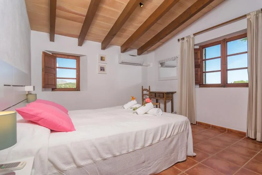 A bright bedroom has a large bed beneath an exposed wooden-beam ceiling.
