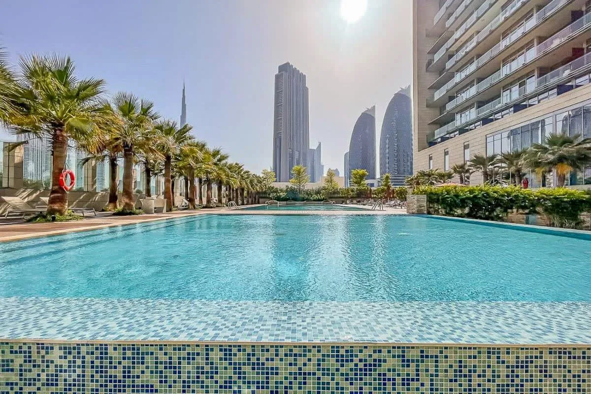 An outdoor swimming pool beside a modern building, with palm trees and city towers in the background.